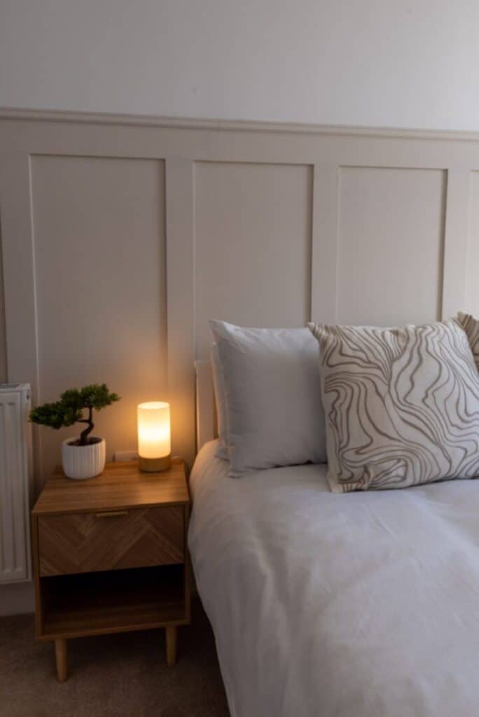 A neatly made bed with white bedding and patterned pillows beside a wooden bedside table holding a small lamp and a potted plant.