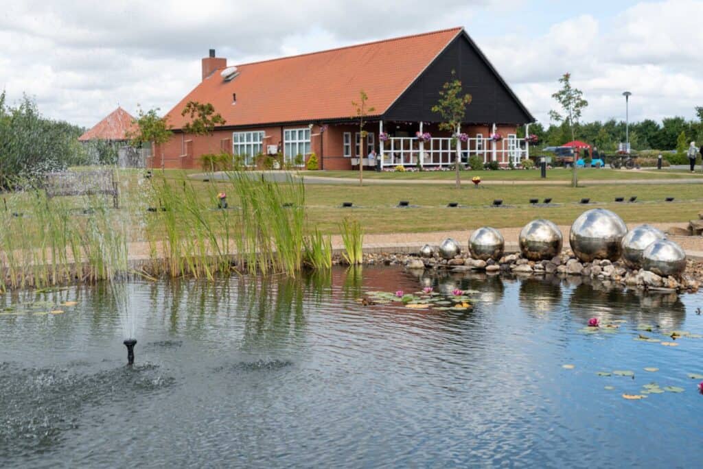 A red-roofed brick building sits behind a pond with water plants, a fountain, and several reflective metal spheres on the shore.