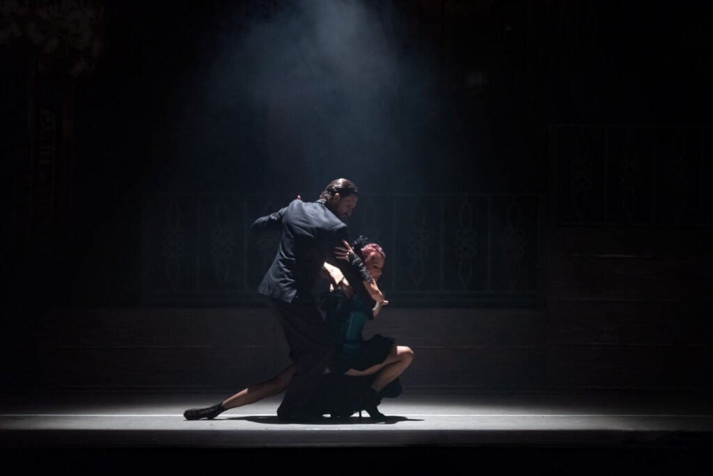 Two dancers perform a dramatic tango onstage under a spotlight, surrounded by shadows and mist.