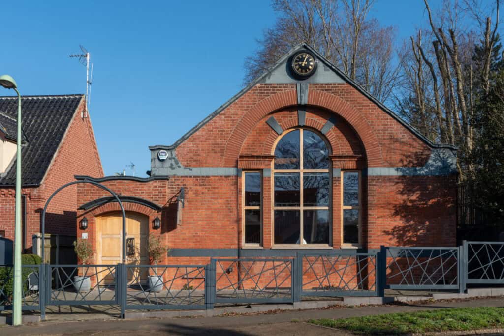A red brick building with a large arched window, clock above the entrance, and a metal gate in front; trees are visible in the background under a clear blue sky.