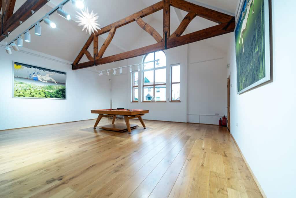 Spacious art gallery room with wooden beams, large arched window, wooden floor, a central table, and artwork displayed on white walls.