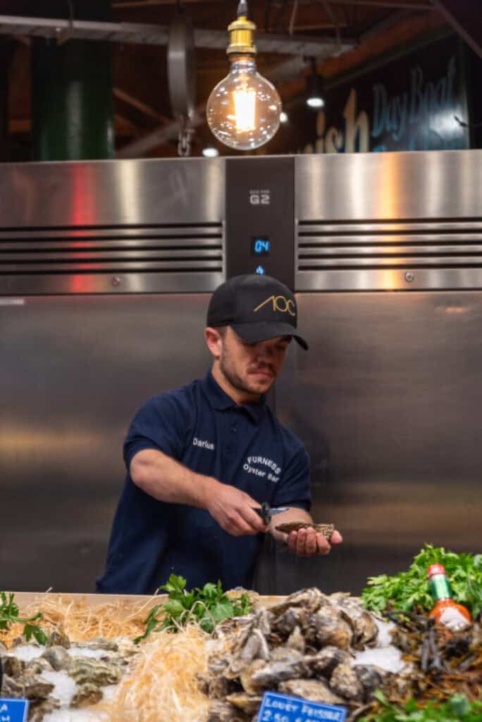 A man in a dark uniform and cap is shucking oysters behind a seafood counter under a hanging lightbulb, with oysters and greens displayed in front of him.