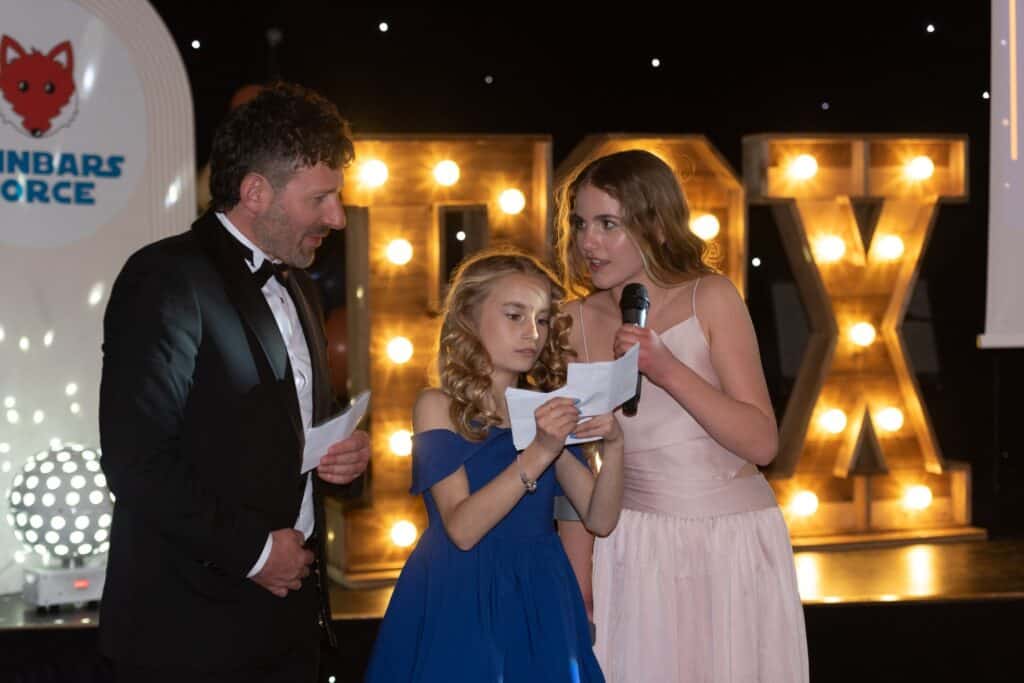 A man in a dinner jacket and two girls in formal dresses are reading from paper on stage with illuminated "FOX" letters in the background.