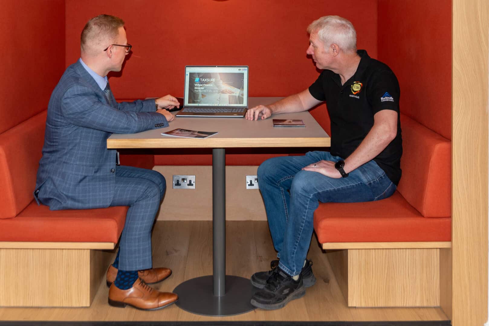 Two men sit opposite each other in an orange booth, looking at a laptop displaying a presentation. Leaflets are on the table.